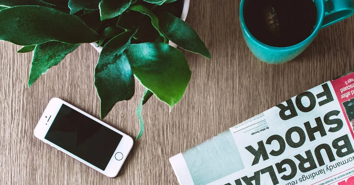 Agencement à plat d'une tasse de café, d'un journal, d'un smartphone et d'une plante sur une table en bois.
